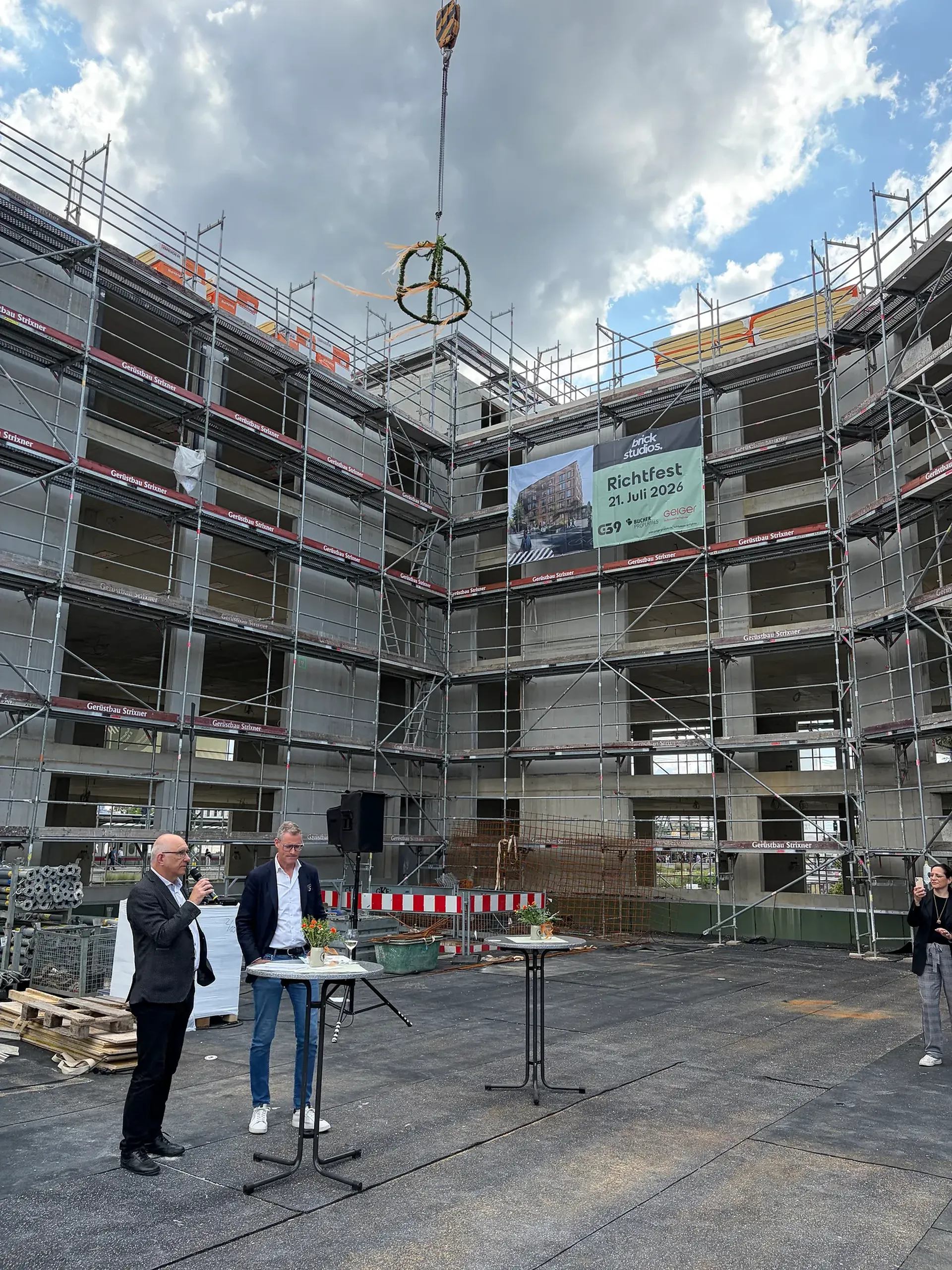 Baustelle mit einem eingerüsteten Gebäude und einem von einem Kran angehobenen Richtkranz. Zwei Männer stehen an Tischen und sprechen zu einer kleinen Gruppe von Gästen. Ein Plakat weist auf das Richtfest am 21. Juli 2026 hin.