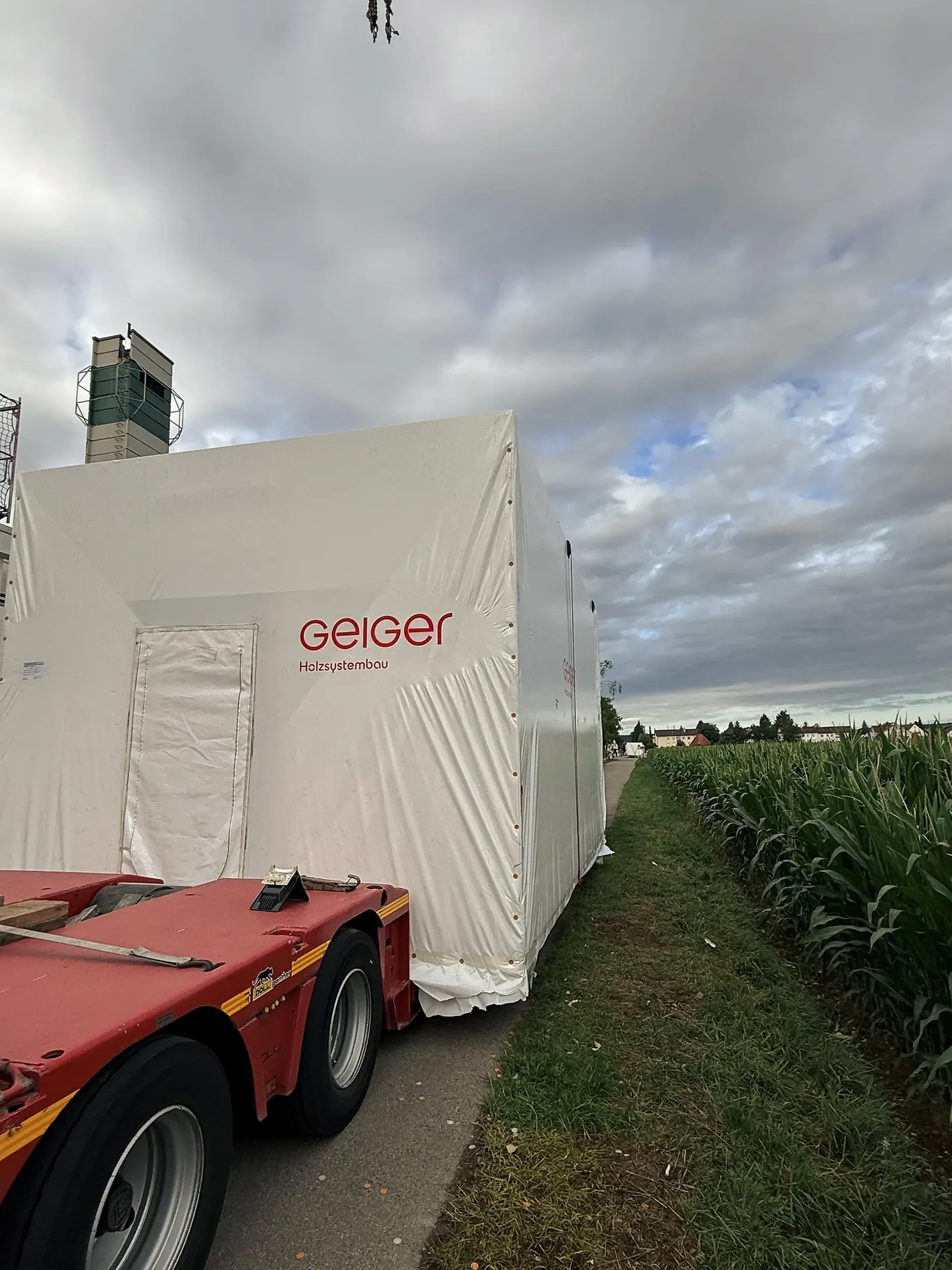 Verpacktes, mit weißer Folie eingepacktes Holzmodul mit der Aufschrift „Geiger Holzsystembau“ auf einem roten Tieflader, neben einem Maisfeld unter einem bewölkten Himmel.