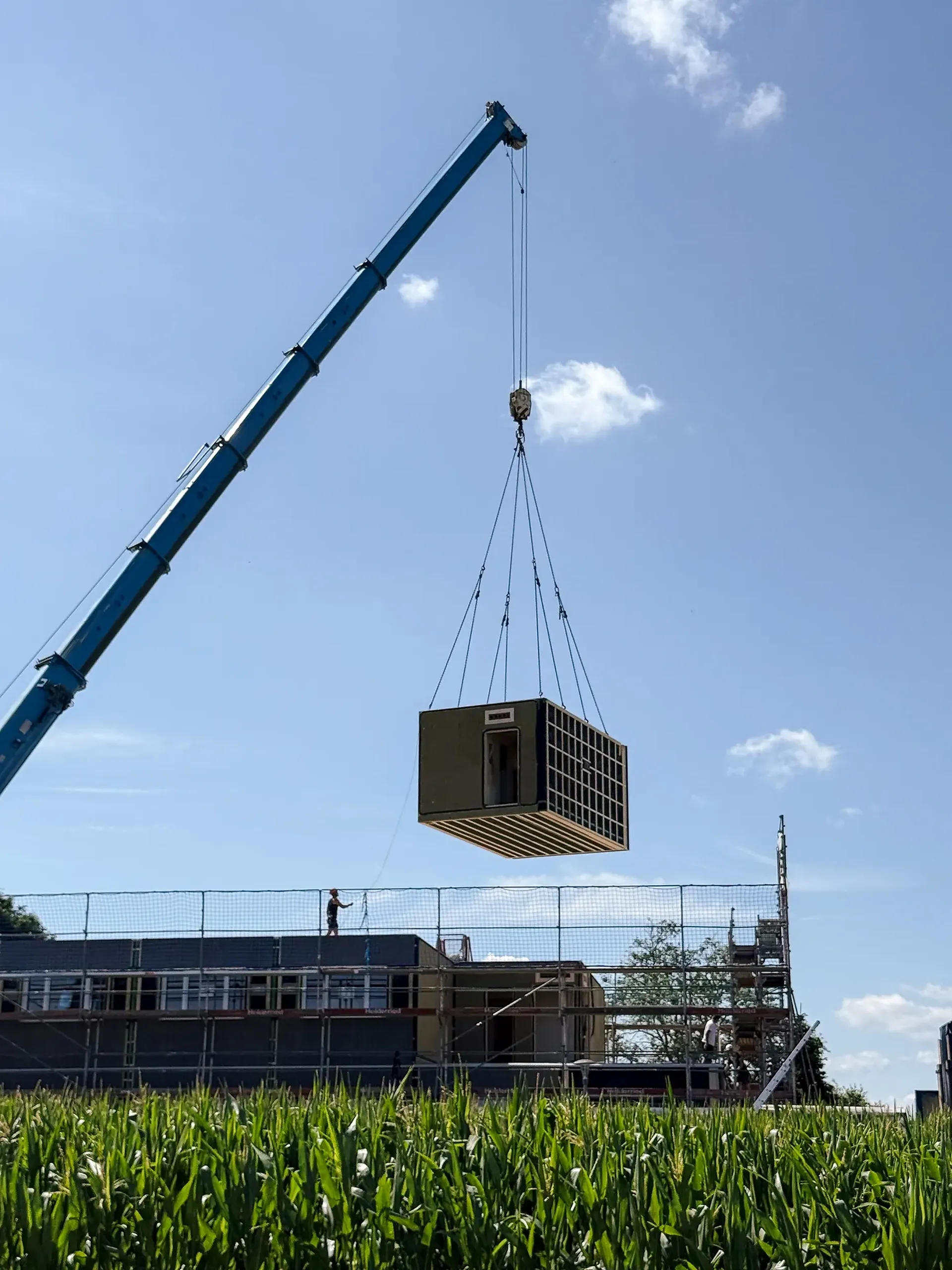 Ein Kran hebt einen modularen Gebäudeabschnitt über eine Baustelle, mit Gerüsten und einem klaren blauen Himmel im Hintergrund.