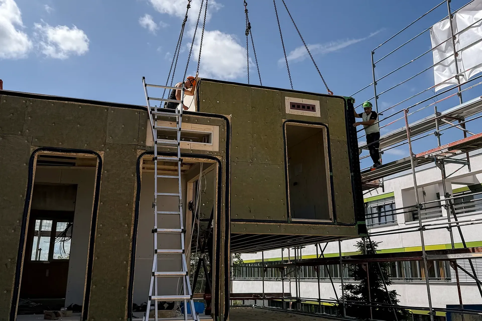 Ein Kran hebt einen modularen Gebäudeteil auf eine Baustelle. Eine Leiter lehnt an der Konstruktion, Arbeiter stehen unter blauem Himmel in der Nähe.