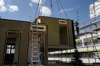 Ein Kran hebt einen modularen Gebäudeteil auf eine Baustelle. Eine Leiter lehnt an der Konstruktion, Arbeiter stehen unter blauem Himmel in der Nähe.