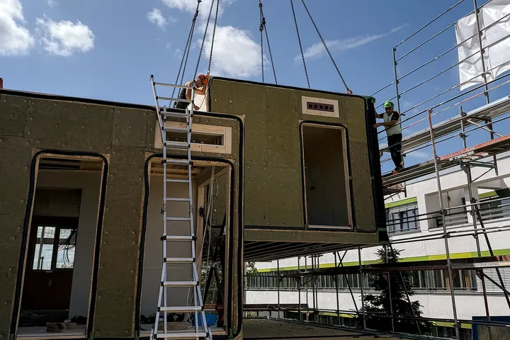 Ein Kran hebt einen modularen Gebäudeteil auf eine Baustelle. Eine Leiter lehnt an der Konstruktion, Arbeiter stehen unter blauem Himmel in der Nähe.
