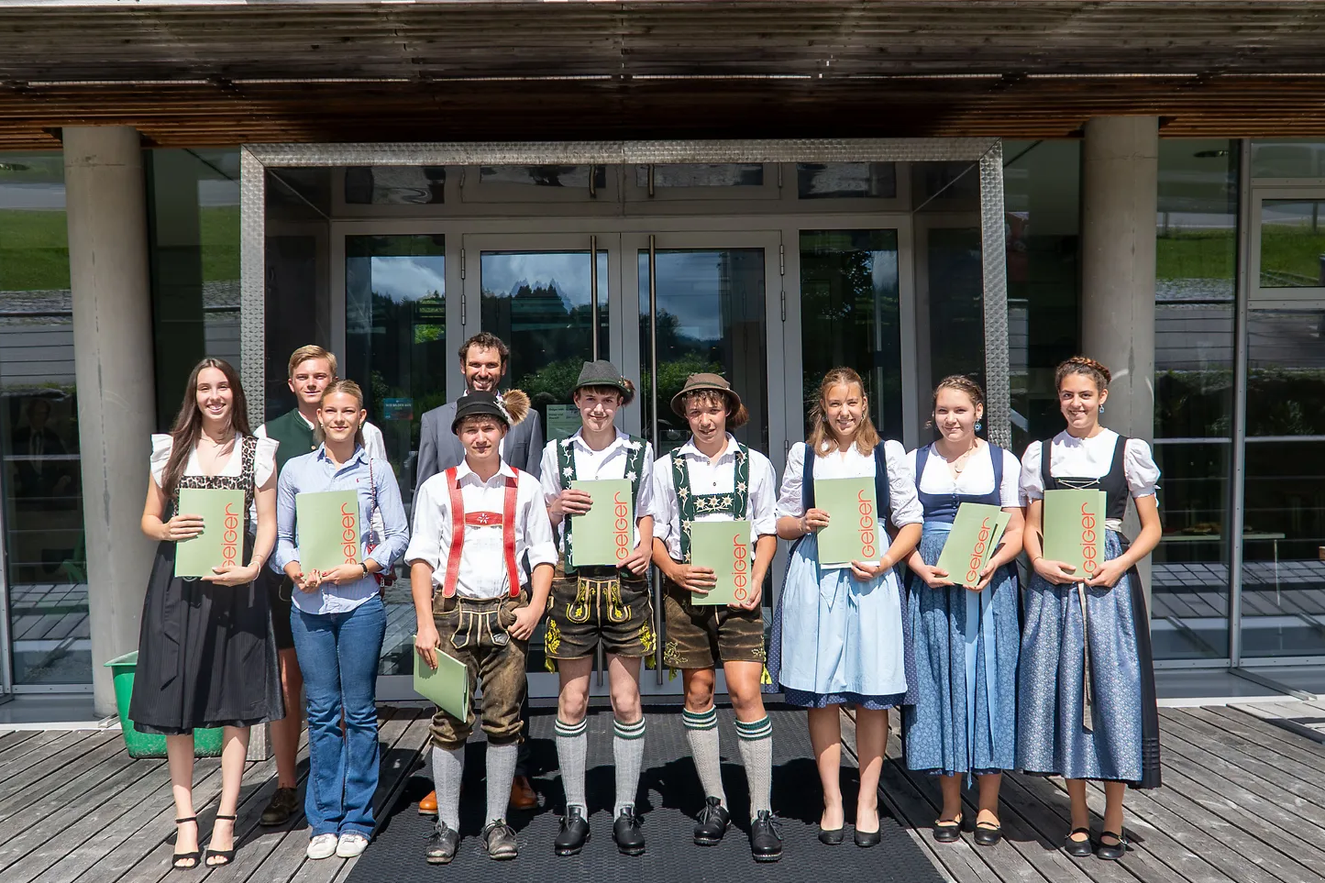 Die Absolventen stehen lächelnd vor einem Gebäude, halten Zertifikate der Geiger Gruppe in den Händen.