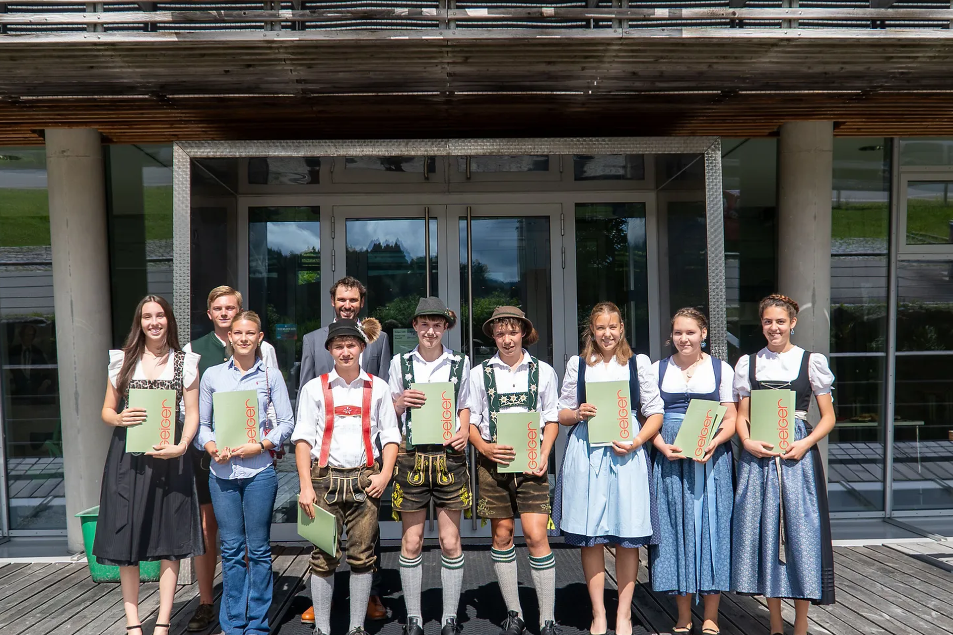 Die Absolventen stehen lächelnd vor einem Gebäude, halten Zertifikate der Geiger Gruppe in den Händen.