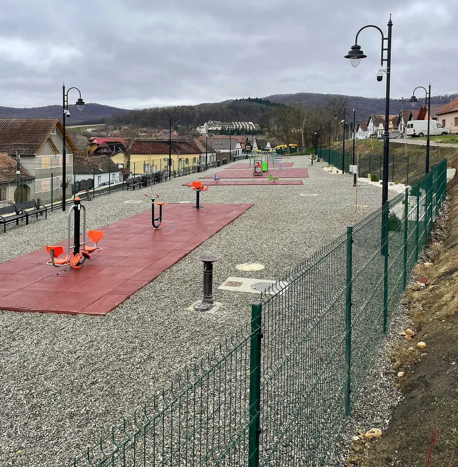 Parc de fitness în aer liber cu echipamente de exerciții pe saltele roșii, înconjurat de alei de pietriș, stâlpi de iluminat și un gard verde într-un cadru rural.