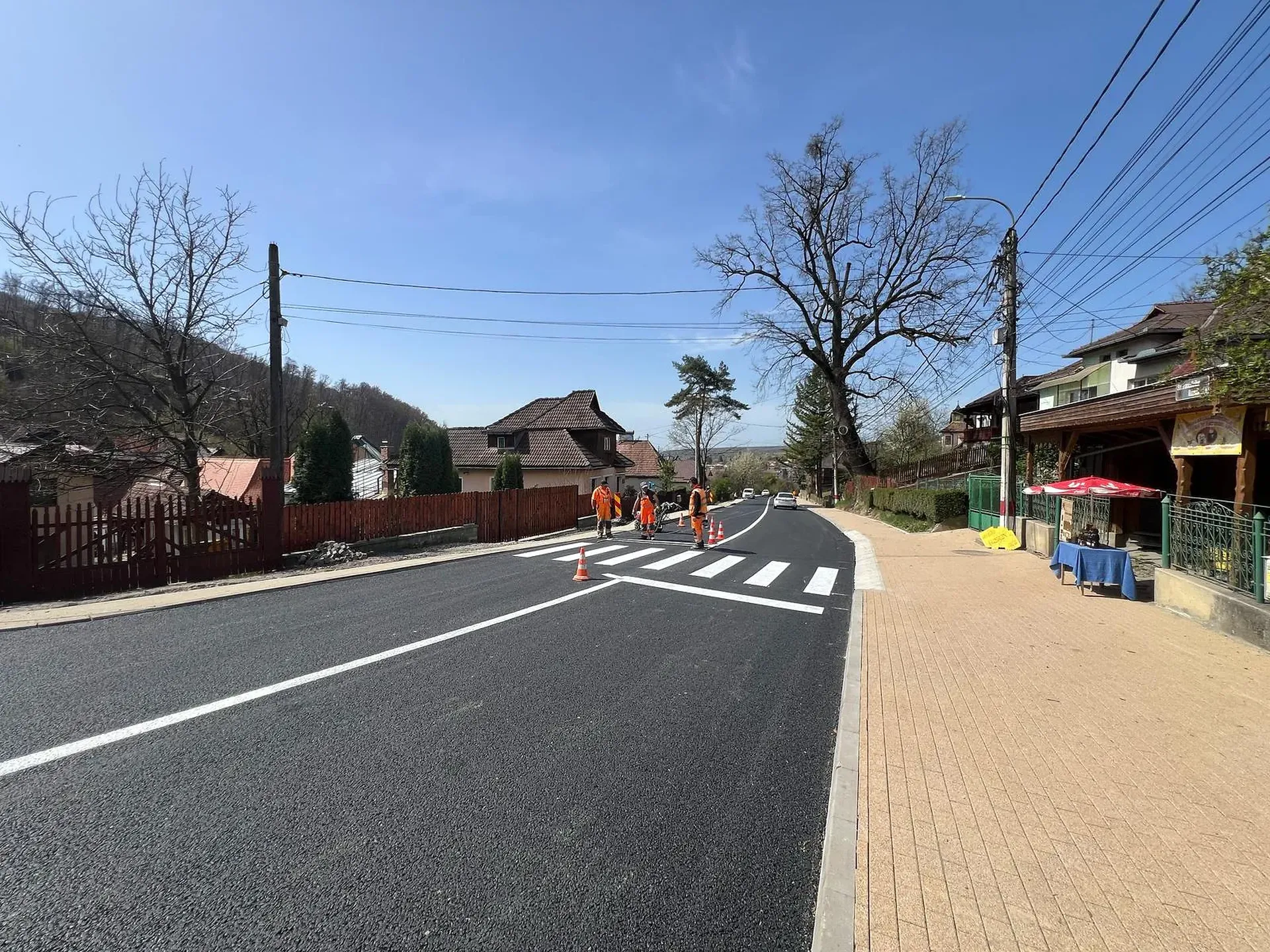 Muncitorii în veste portocalii pictează liniile de trecere pentru pietoni pe un drum proaspăt asfaltat printr-o zonă rezidențială, sub un cer senin.