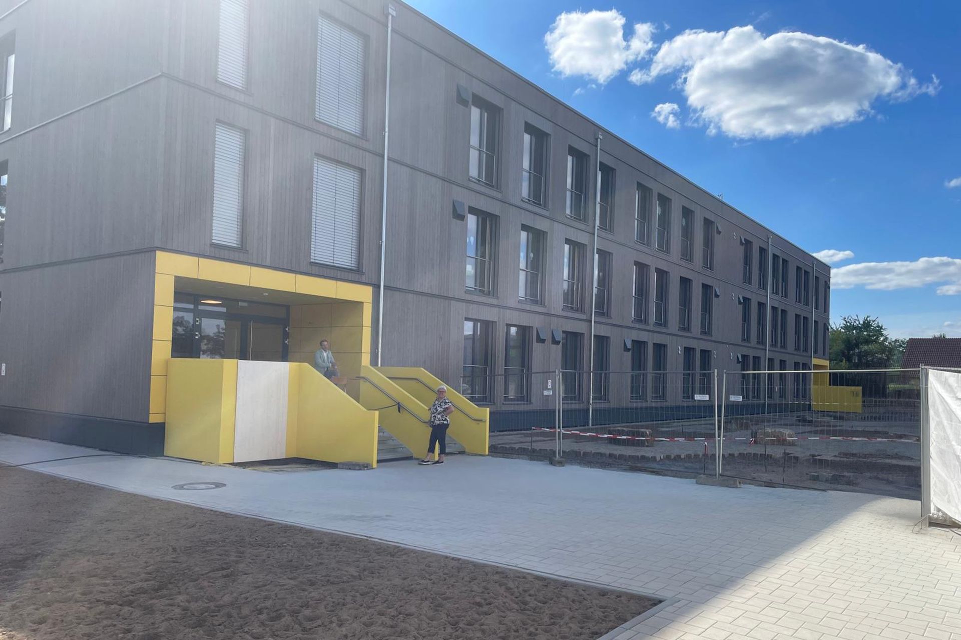 Ein modernes, dreistöckiges Gebäude mit großen Fenstern, gelben Akzenten und einem eingezäunten Bereich unter einem klaren blauen Himmel mit Wolken