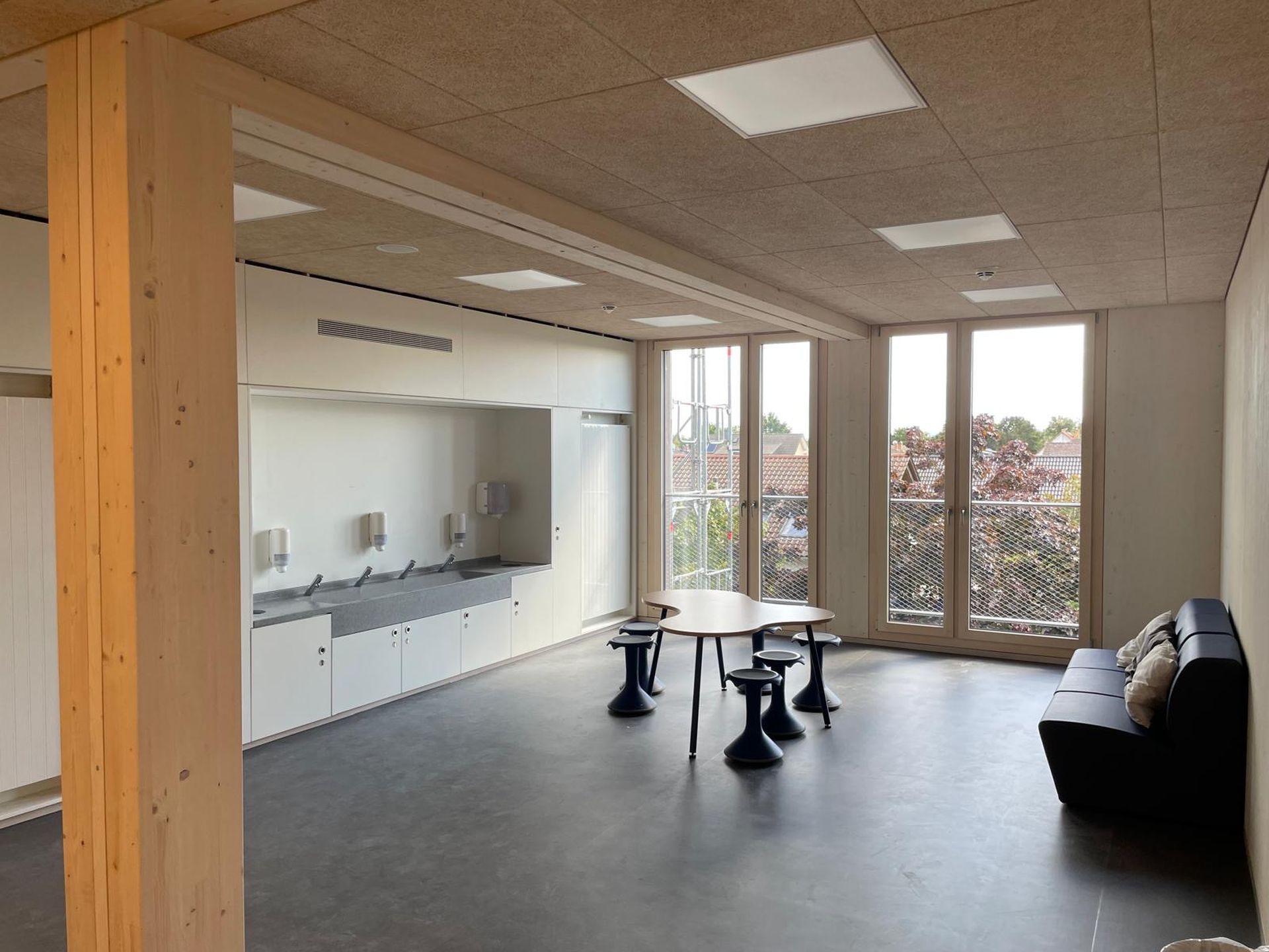 Ein moderner, minimalistischer Raum mit großen Fenstern, einem schwarzen Sofa, einem Holztisch mit Hockern und einer Reihe von Waschbecken entlang der Wand.