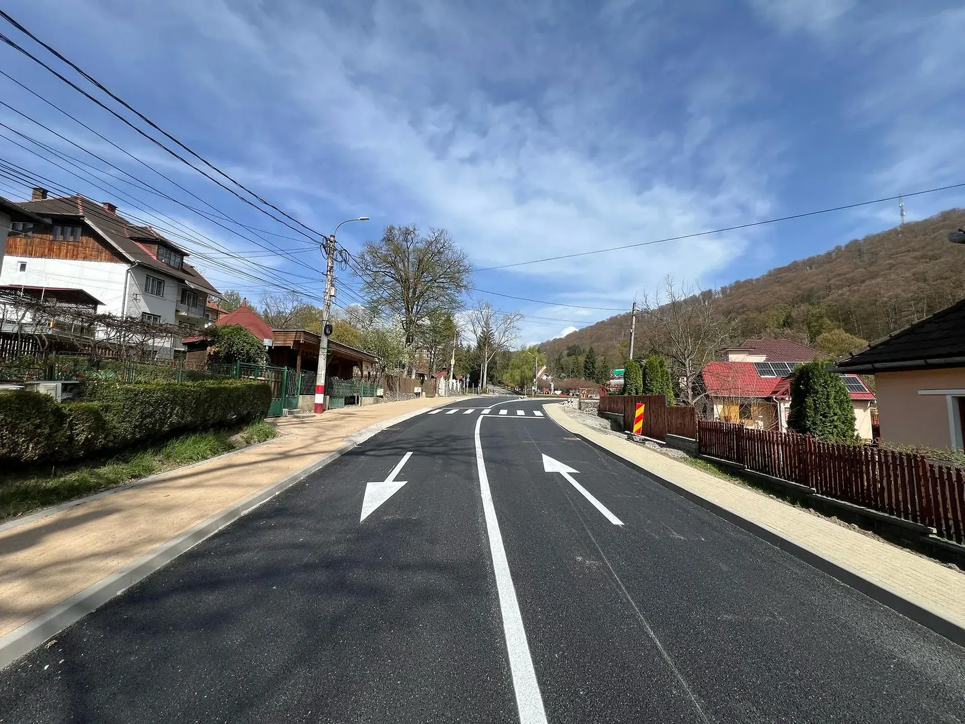 Un drum proaspăt pavat cu marcaje clare de bandă, flancat de case și vegetație sub un cer albastru parțial noros într-o zonă deluroasă.