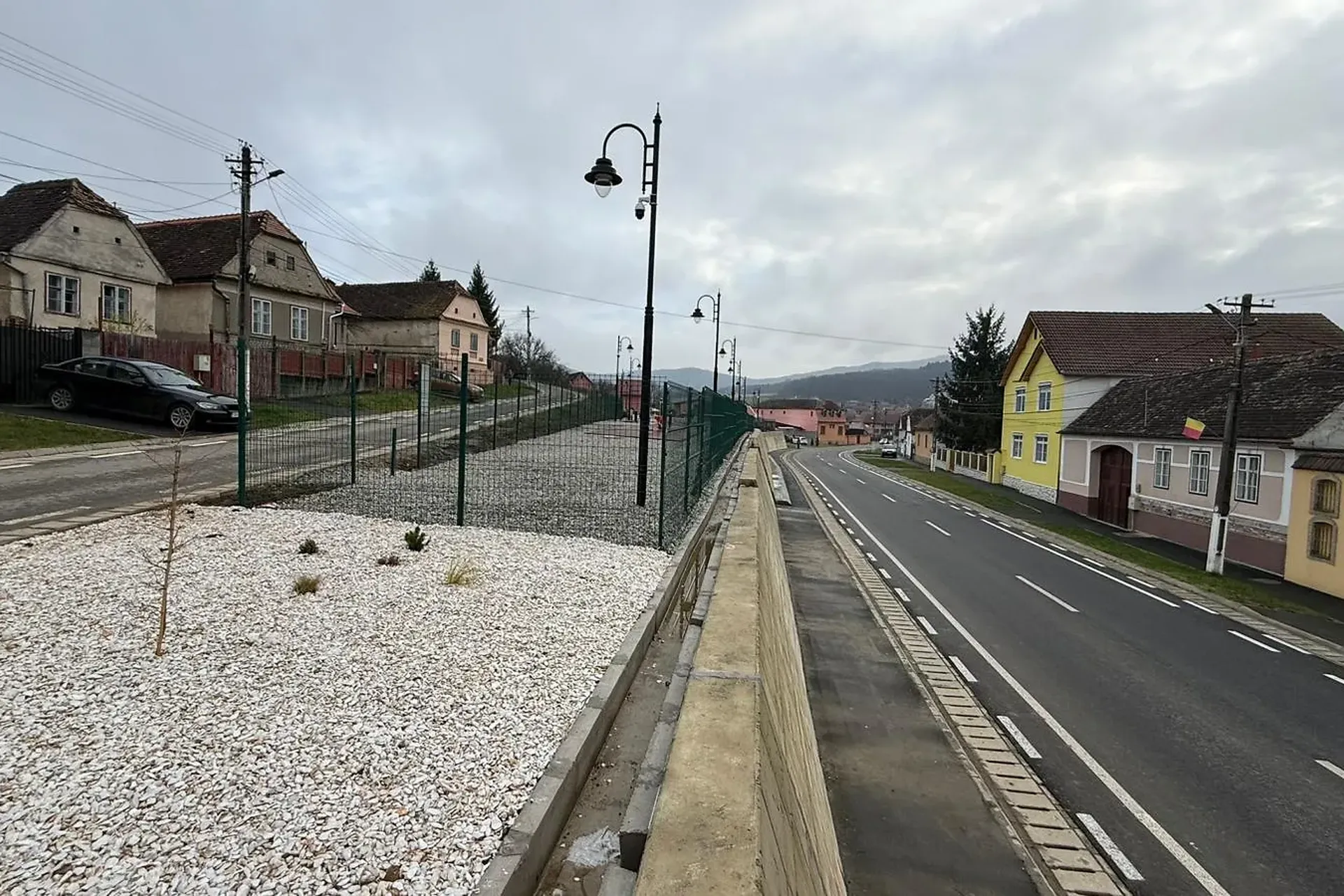 Vedere aeriană a unui pod rural care traversează un râu, înconjurat de câmpuri cu iarbă și copaci, cu un drum care duce către o mică așezare în depărtare. ...