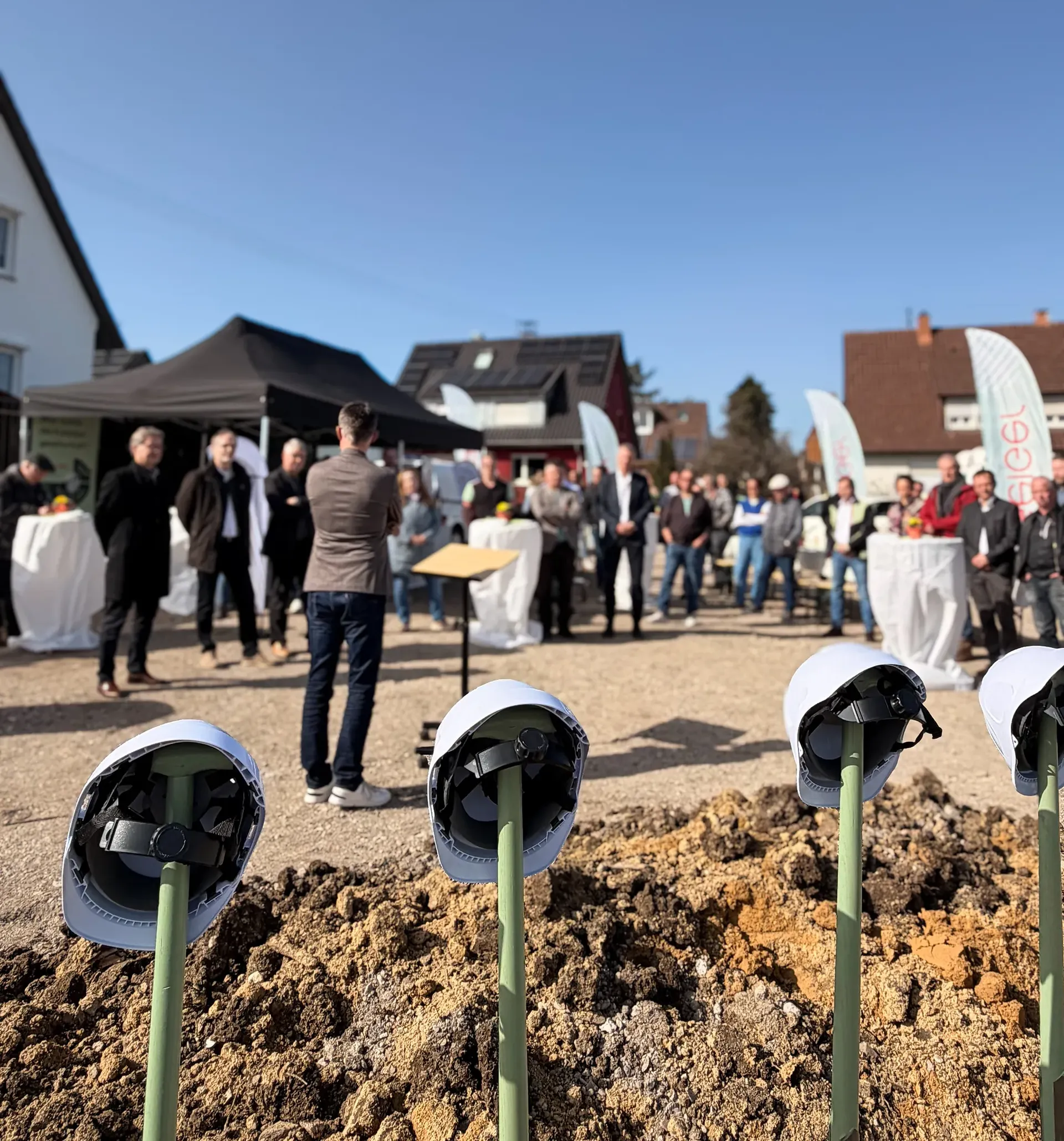Eine Gruppe von Menschen versammelte sich im Freien auf einer Baustelle, mit Helmen auf Ständern im Vordergrund, unter einem klaren blauen Himmel.