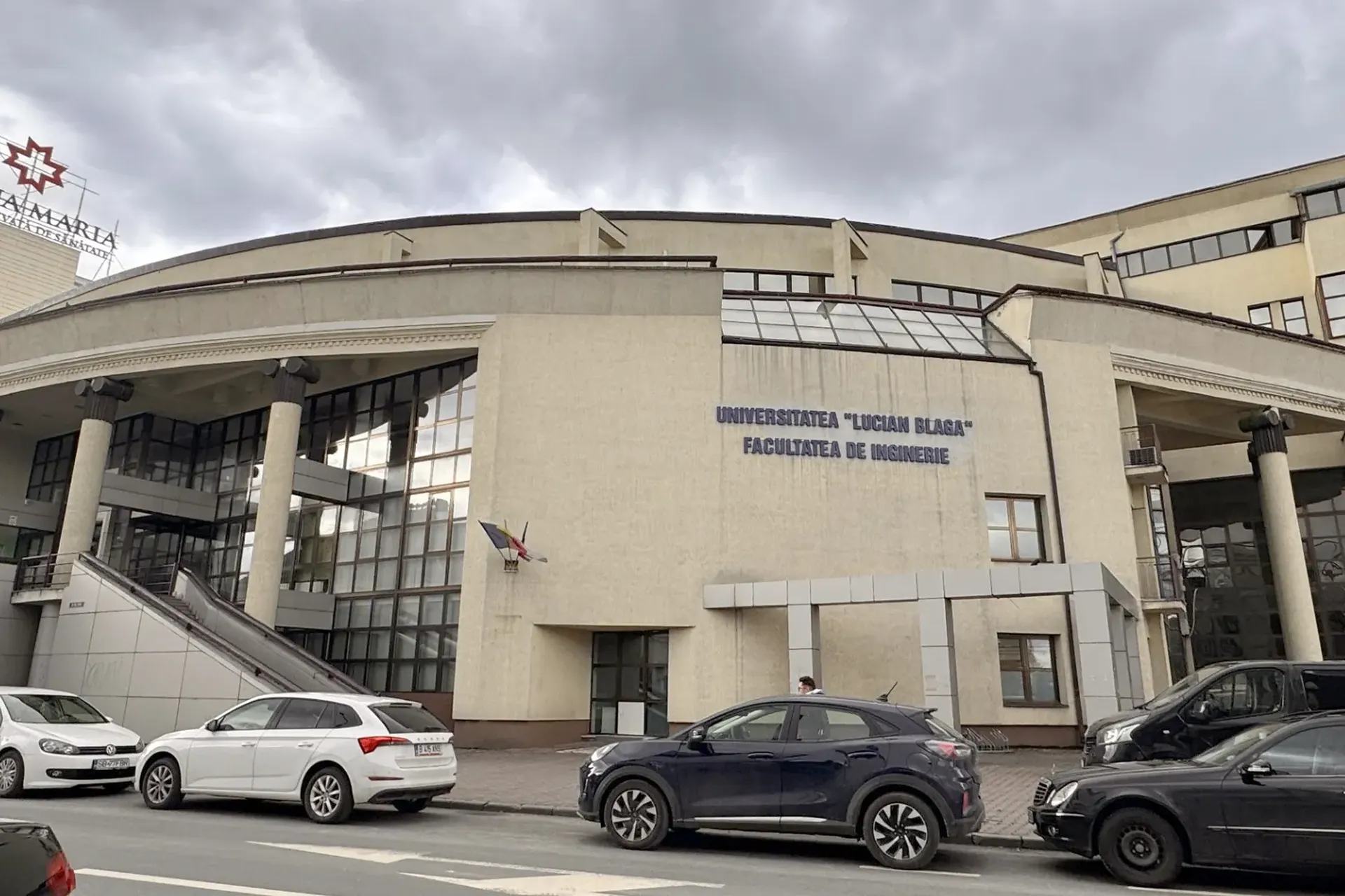 Vedere stradală a clădirii Facultății de Inginerie „Universitatea Lucian Blaga”, cu mașini parcate și un cer înnorat deasupra.