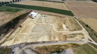 Luftaufnahme einer Baustelle mit Baumaschinen, die auf einer großen, geräumten Fläche arbeiten, umgeben von grünen Feldern und Straßen.