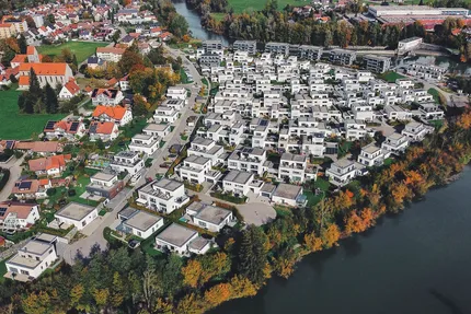 Luftaufnahme des Wohnprojekts "Am Illerbogen daheim" mit Fokus auf die moderne Architektur der Wohnhäuser, die Nähe zur Flusslandschaft und die Integration in die Natur. Gebäude mit Photovoltaikanlagen sind im Vordergrund sichtbar.