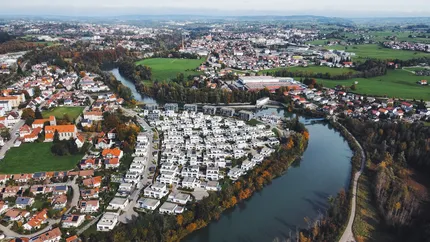 Panorama-Luftaufnahme des Wohnquartiers "Am Illerbogen daheim". Die moderne Siedlung fügt sich harmonisch in die Flusslandschaft ein, umgeben von Dörfern, Wiesen und Feldern. Im Hintergrund ein Berg- und Dorfpanorama.