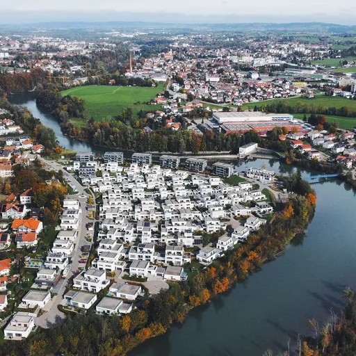 Panorama-Luftaufnahme des Wohnquartiers "Am Illerbogen daheim". Die moderne Siedlung fügt sich harmonisch in die Flusslandschaft ein, umgeben von Dörfern, Wiesen und Feldern. Im Hintergrund ein Berg- und Dorfpanorama.