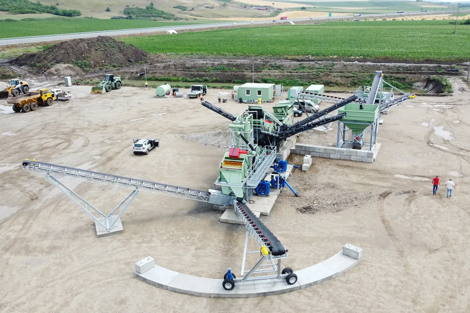 Vedere aeriană a unui sit industrial care arată utilaje, benzi transportoare, vehicule și muncitori pe un teren de pământ, cu câmpuri verzi în fundal.