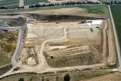 Luftaufnahme einer Baustelle mit Baumaschinen, Erdwegen und geräumten Flächen, umgeben von Feldern und einer Straße in ländlicher Umgebung.