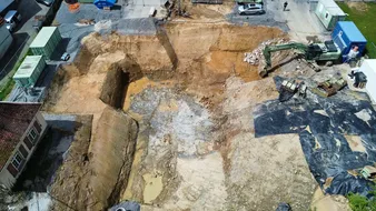 Luftaufnahme einer großflächigen Baugrube mit Erdbewegungsmaschinen, umgeben von Containern, Fahrzeugen und einem Gebäude am Rand des Geländes.