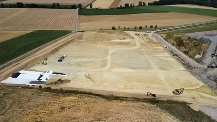 Luftaufnahme einer Baustelle mit Fahrzeugen und Baumaschinen auf einer großflächig planierten Erdfläche, umgeben von Feldern und vereinzelten Grünflächen.