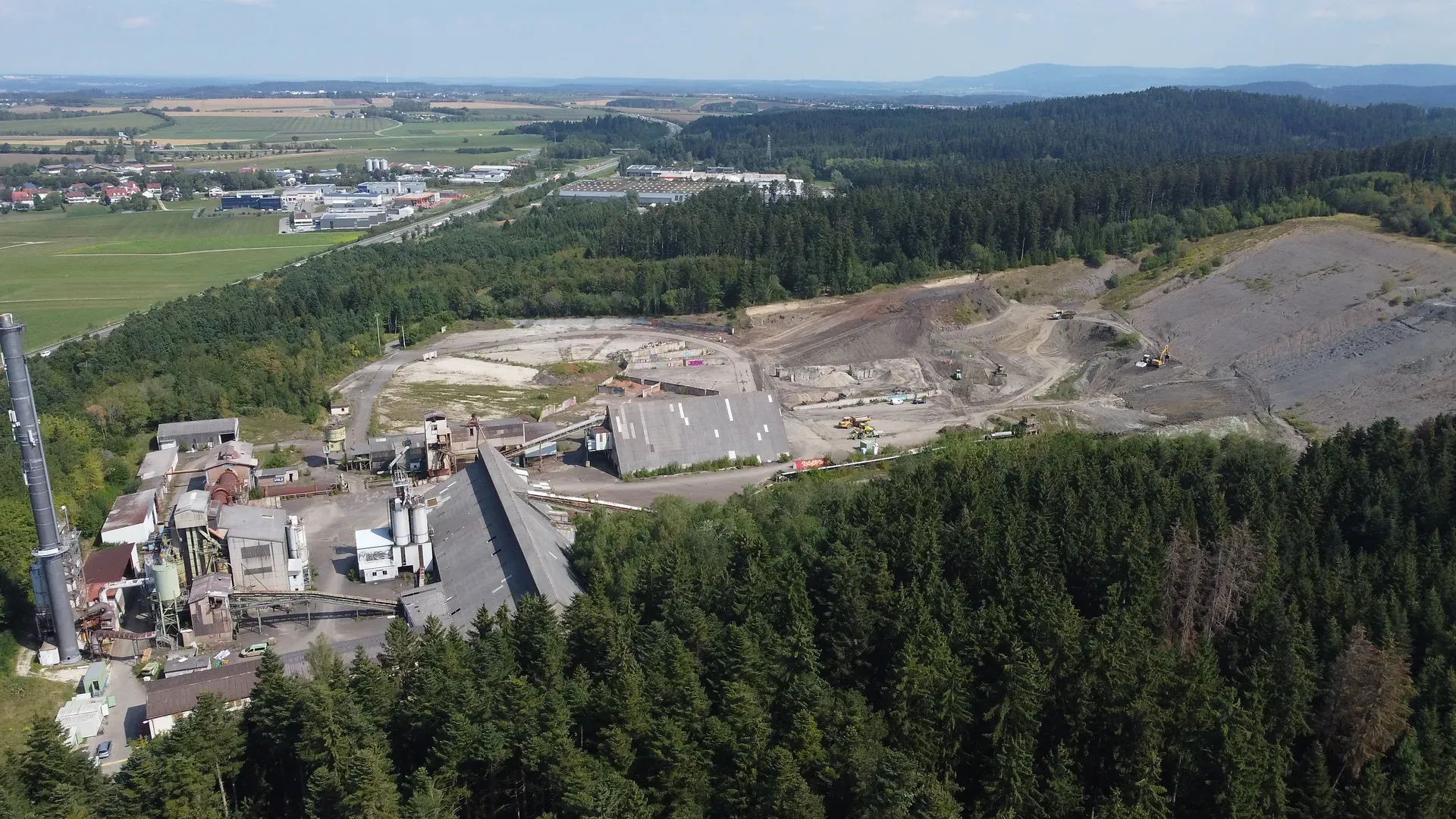 Luftaufnahme eines Industriekomplexes in der Nähe eines Waldes, mit umliegenden Feldern, Gebäuden und entfernten Hügeln unter einem blauen Himmel.