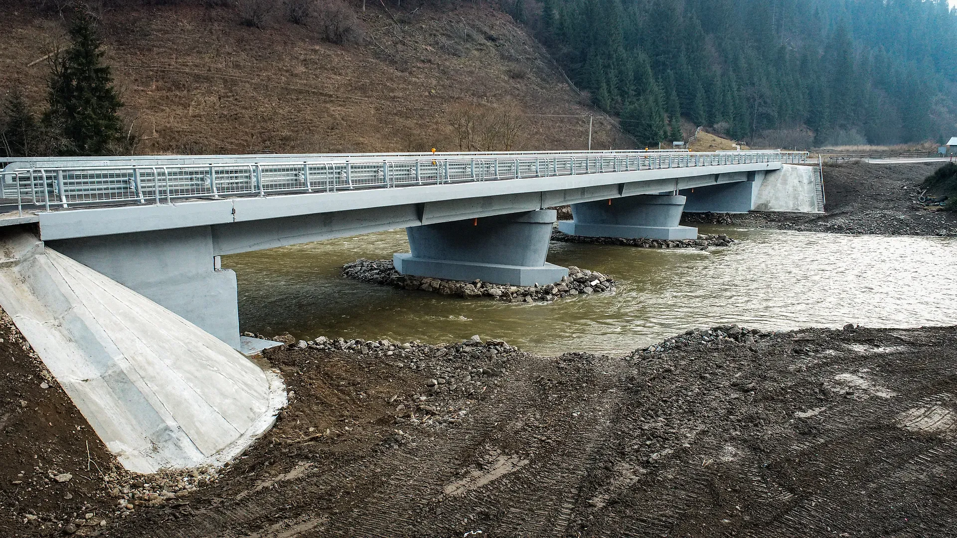 Un pod de beton traversează un râu curgător, înconjurat de copaci și de malurile stâncoase ale râului.