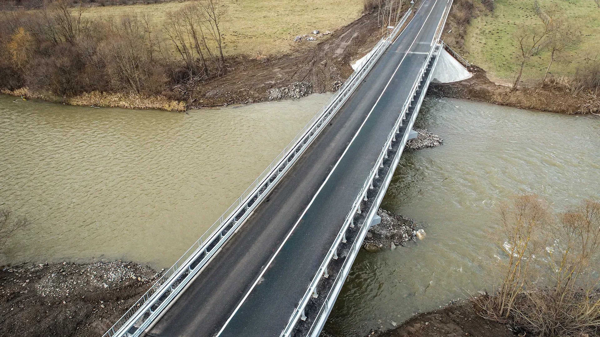 Vedere aeriană a unui pod cu două benzi care traversează un râu noroios, înconjurat de copaci fără frunze și câmpuri cu iarbă.