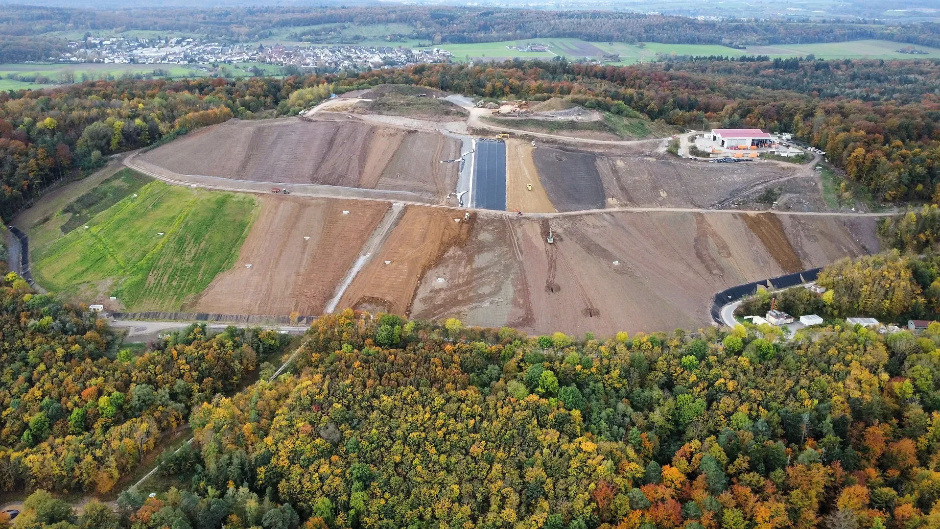 Luftaufnahme einer großen Baustelle, umgeben von herbstlichem Wald, mit Feldern und einer entfernten Stadt im Hintergrund.