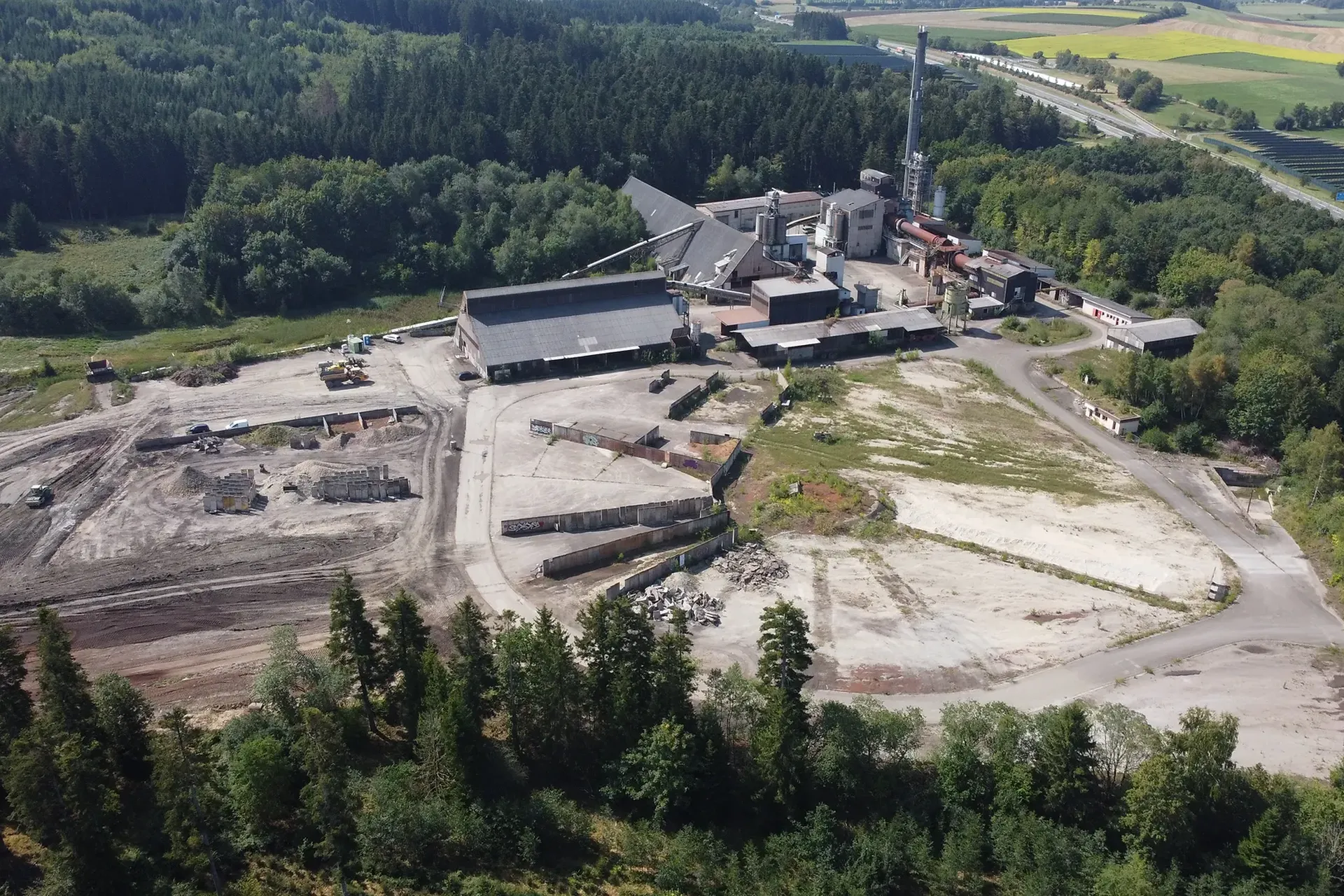 Luftaufnahme einer Industrieanlage, umgeben von Wald und Feldern, mit Gebäuden, Straßen und gerodeten Flächen