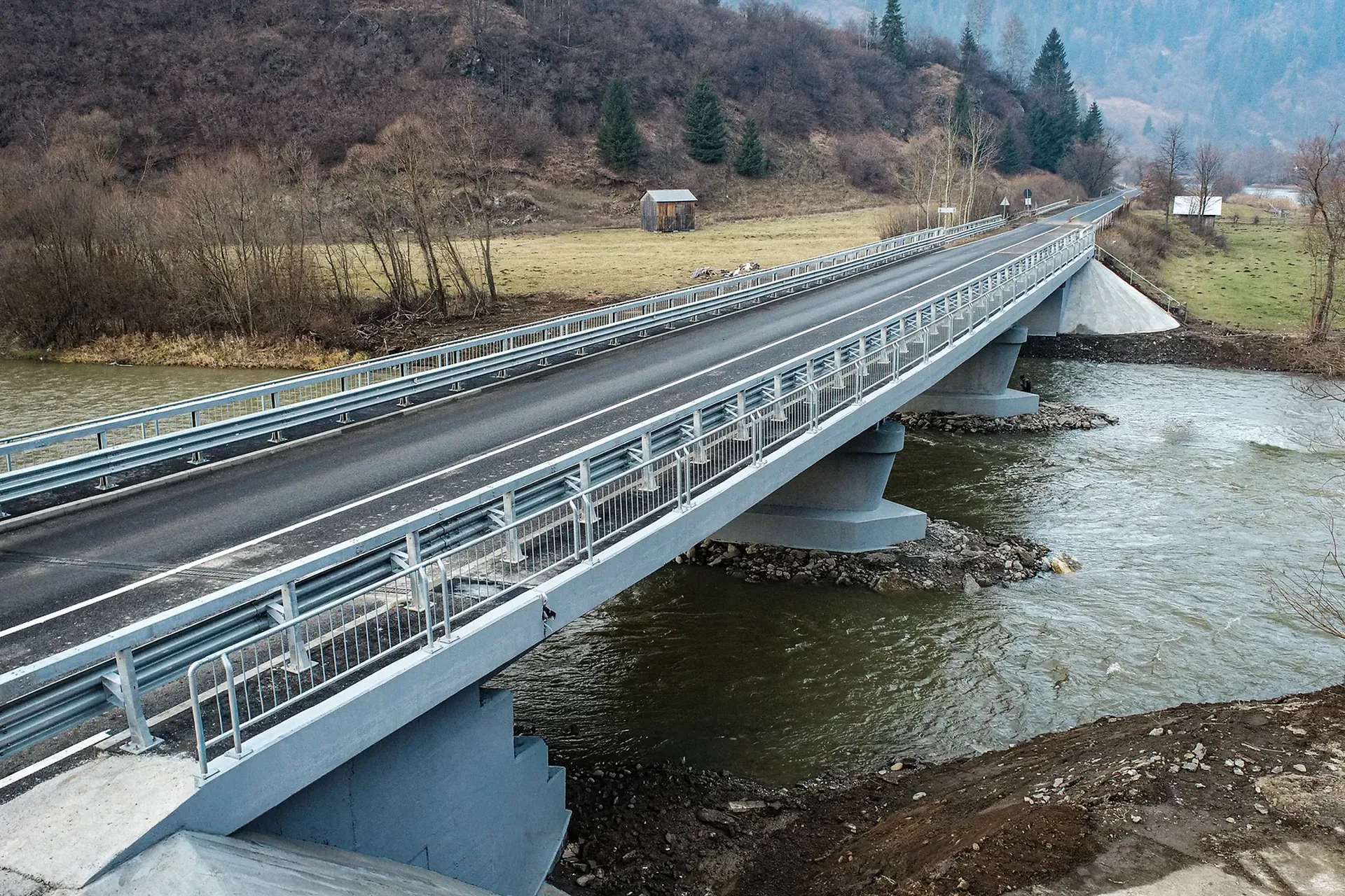 Un pod lung de beton cu balustrade metalice se întinde peste un râu curgător, înconjurat de un peisaj rural cu copaci și construcții mici.