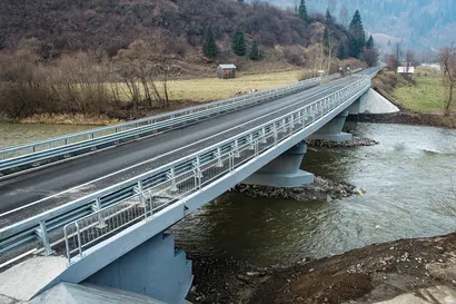 Un pod lung de beton cu balustrade metalice se întinde peste un râu curgător, înconjurat de un peisaj rural cu copaci și construcții mici.