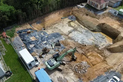 Luftaufnahme einer Baustelle mit einer großen Baugrube, einem Bagger und weiterer Baustellenausrüstung. In der Nähe sind ein kleines Haus und Bäume zu sehen.