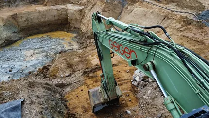 Bagger mit der Aufschrift „Geiger“ am Ausleger, der auf einer großen, felsigen Baustelle mit freiliegender Erde und einem flachen Wasserbecken gräbt.