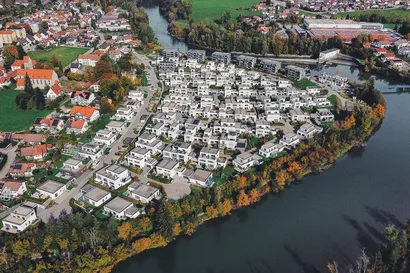 Panorama-Luftaufnahme des Wohnquartiers "Am Illerbogen daheim". Die moderne Siedlung fügt sich harmonisch in die Flusslandschaft ein, umgeben von Dörfern, Wiesen und Feldern.