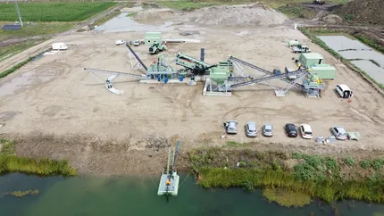 Vedere aeriană a unui sit industrial cu utilaje, mașini parcate și un mic corp de apă în prim-plan.