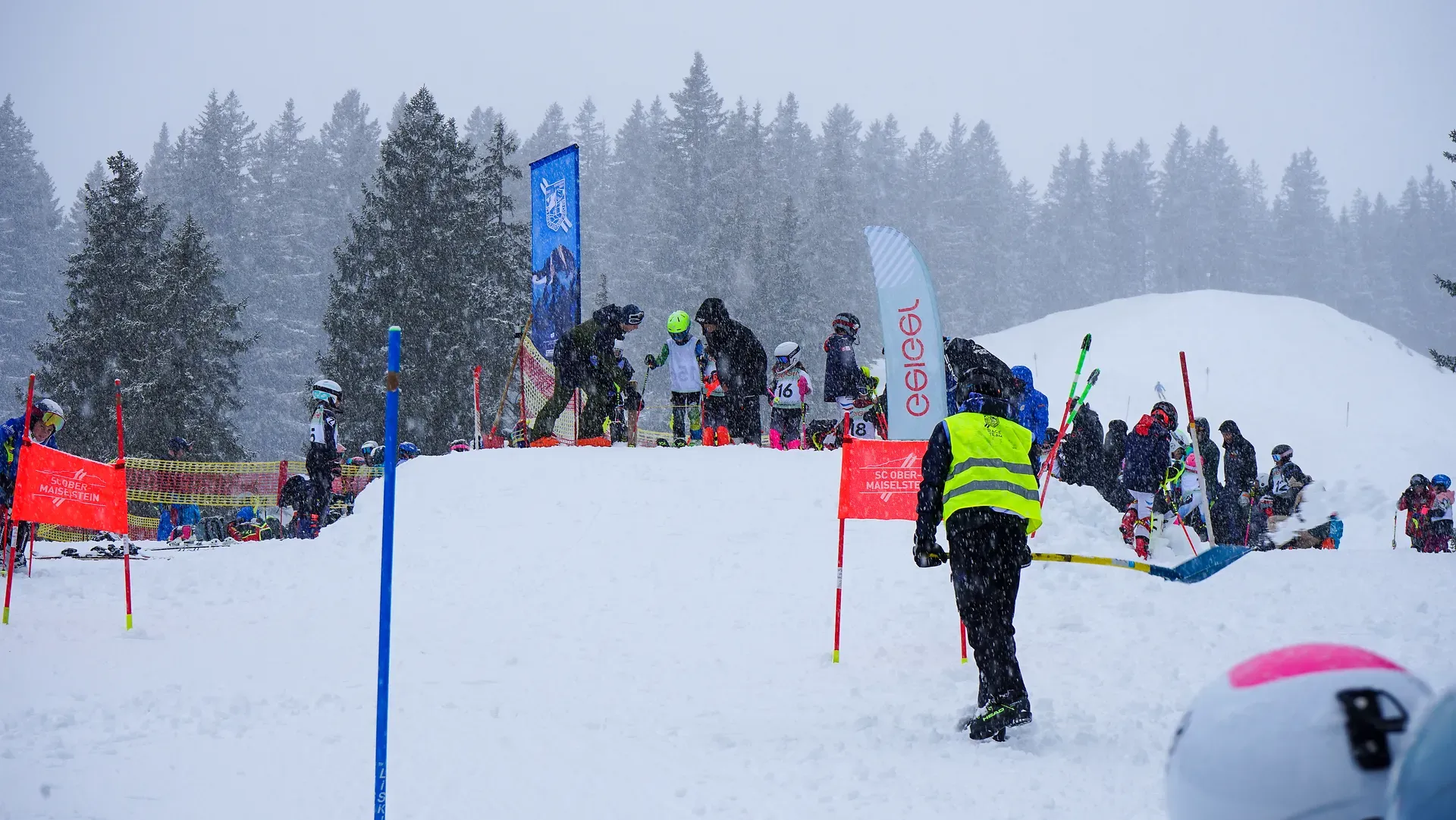 Skifahrer und Snowboarder versammeln sich an einem verschneiten Hang, umgeben von Fahnen und Bäumen, während eine Person in einer gelben Weste hilft. Sanft fällt Schnee.