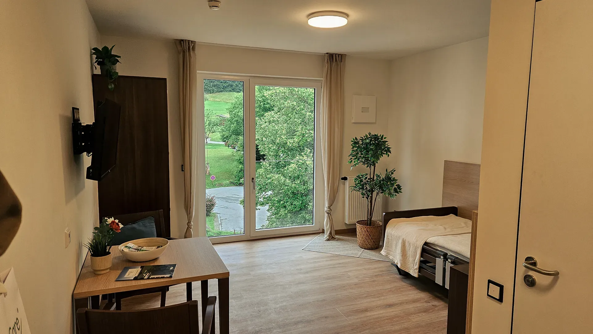 Ein gemütliches Zimmer des Pflegezentrums Lechbruck mit einem Einzelbett, einem kleinen Esstisch und einem großen Fenster mit Blick ins Grüne, mit Holzboden und Topfpflanzen.