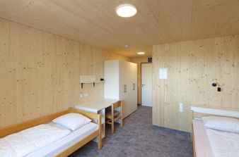Ein Zimmer mit Holzwänden, grauen Teppich, zwei Betten mit Kopfkissen und Bettdecke, ein Schrank und ein Schreibtisch.