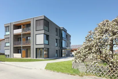 Dreigeschossiges Mehrfamilienhaus in Würfelform aus Holzmodulen mit Balkonen, rechts unten im Bild ein blühender Busch.