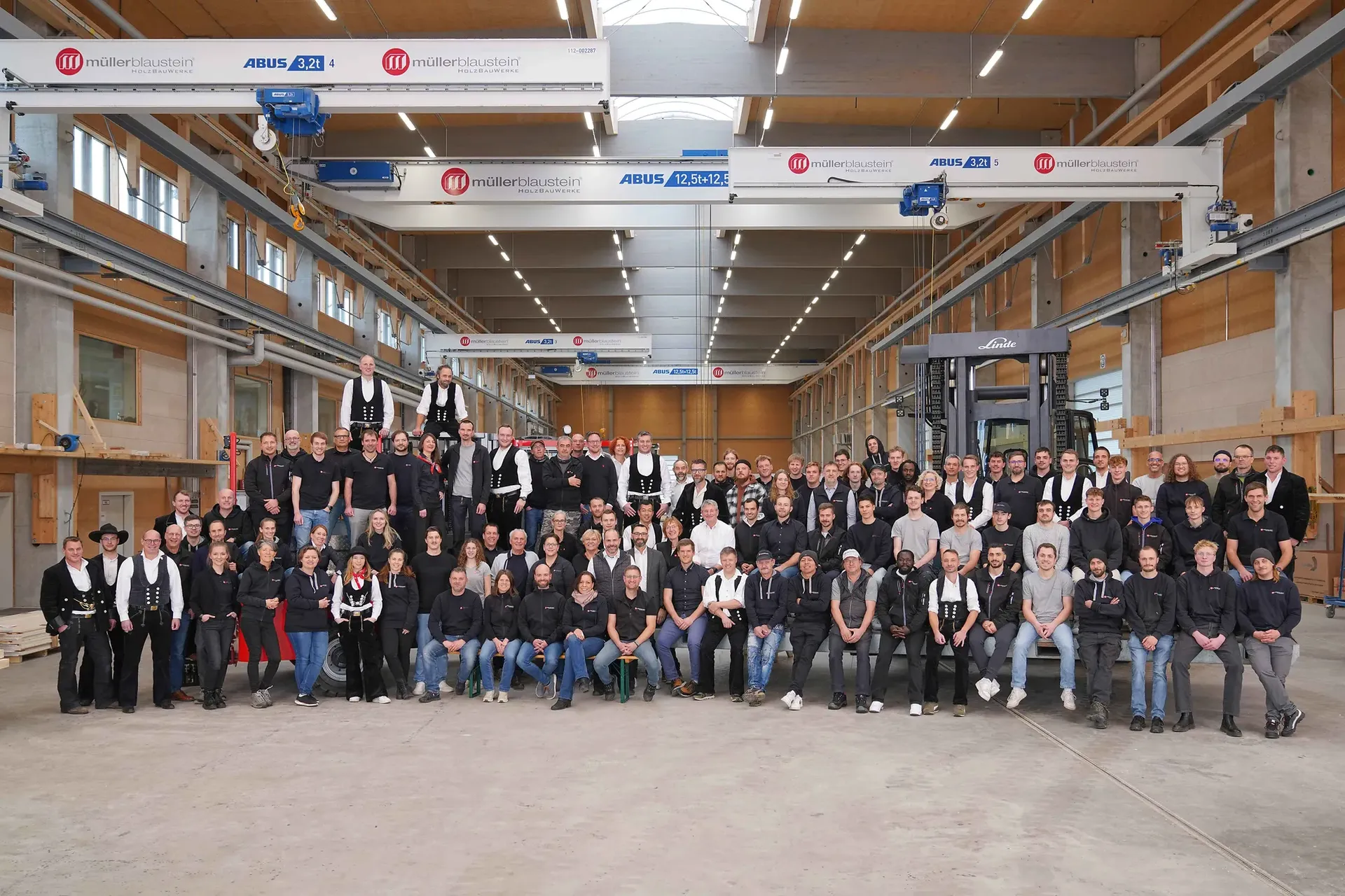 Eine große Gruppe von Menschen posiert in einem geräumigen Industrie­lagerhaus mit Holzbalken, hohen Decken und Laufkränen