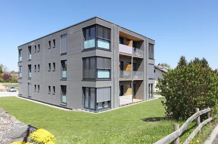Dreigeschossiges Mehrfamilienhaus in Würfelform aus Holzmodulen mit Balkonen und grünem Garten.
