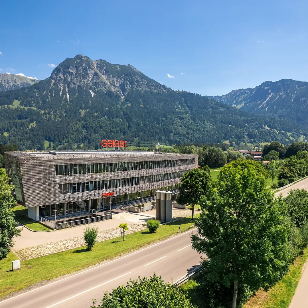 Luftaufnahme der Hauptzentrale der Geiger Gruppe in Oberstdorf, eingebettet in eine Landschaft mit grünen Wiesen und Bergen im Hintergrund.