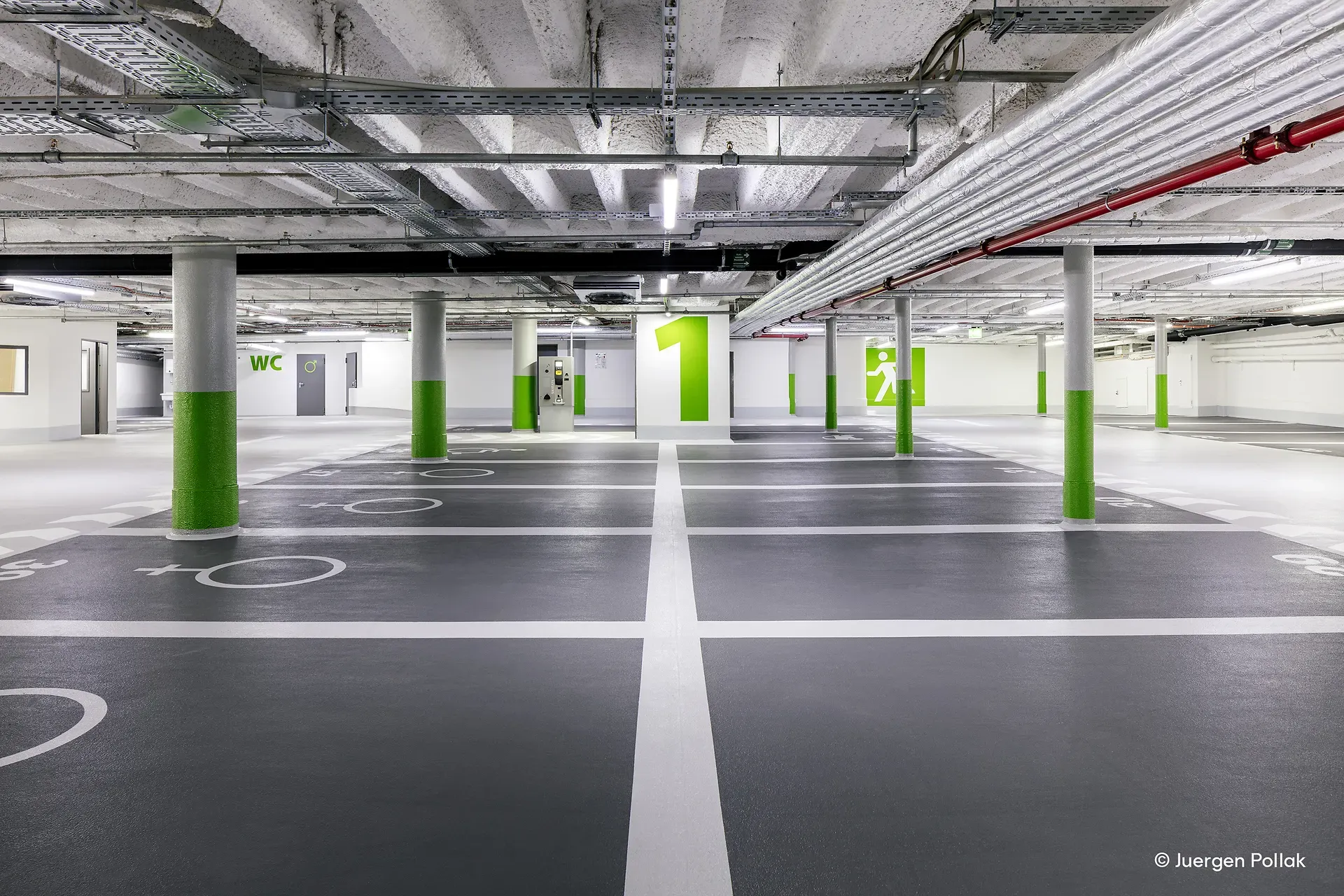 Leeres unterirdisches Parkhaus mit grauen Böden, grünen und weißen Säulen, Deckenrohren und markierten Parkplätzen. Eine große Zahl „1“ an einer Wand.