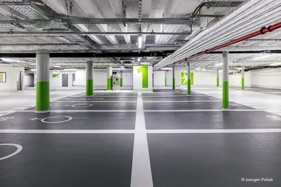 Leeres unterirdisches Parkhaus mit grauen Böden, grünen und weißen Säulen, Deckenrohren und markierten Parkplätzen. Eine große Zahl „1“ an einer Wand.