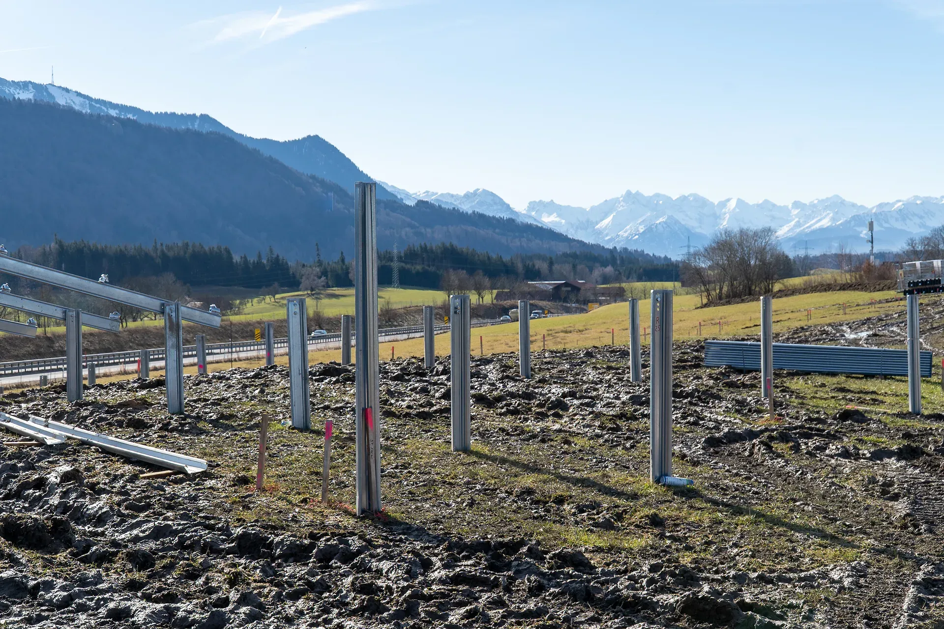 Metallpfosten stehen auf eine Baustelle, im Hintergrund sind Berge und grüne Felder unter einem klaren blauen Himmel zu sehen.