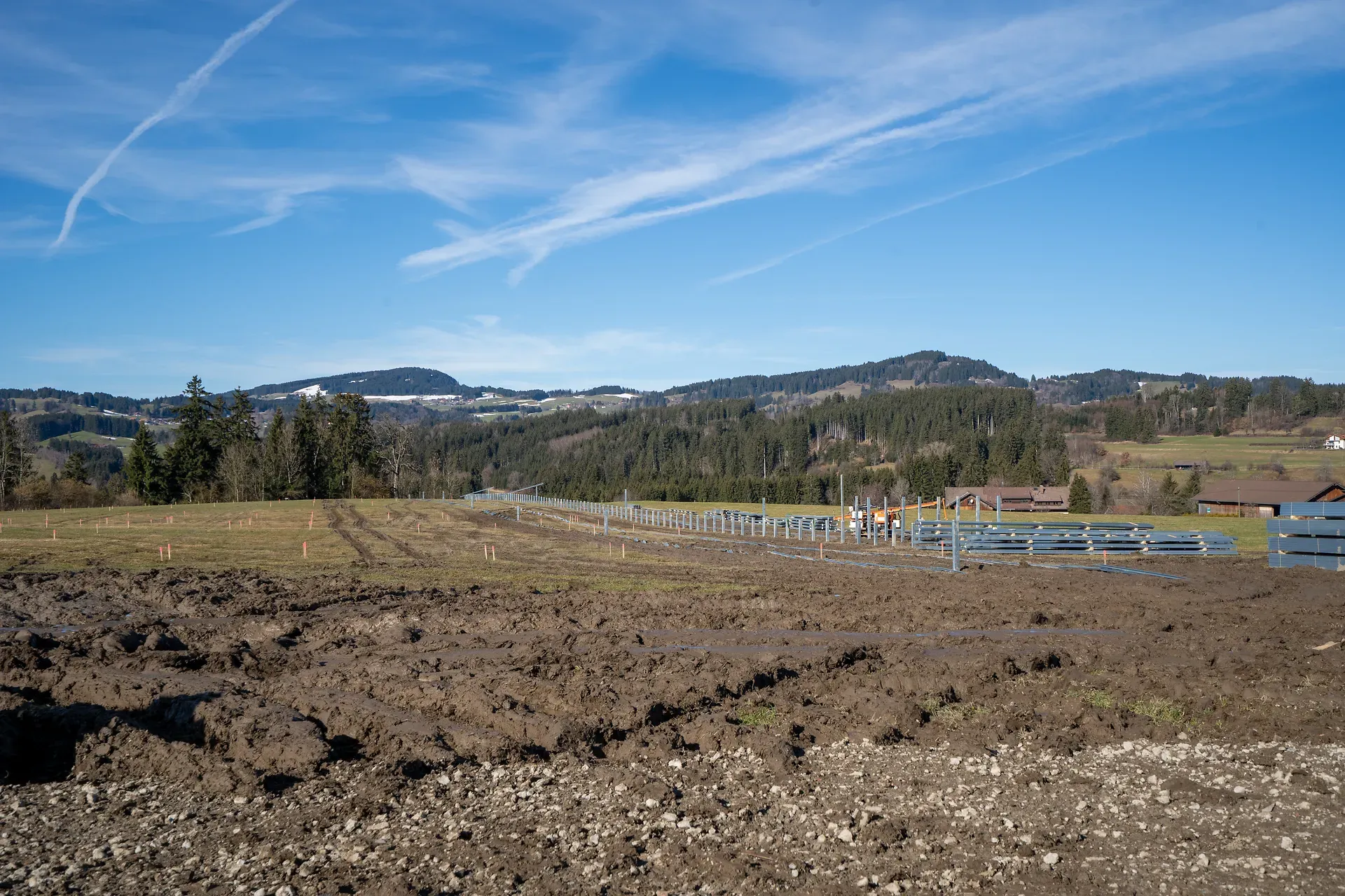 Eine ländliche Landschaft mit einem schlammigen Feld, entfernten Hügeln, verstreuten Bäumen und blauem Himmel mit feinen Wolken. Baumaterialien sind sichtbar.