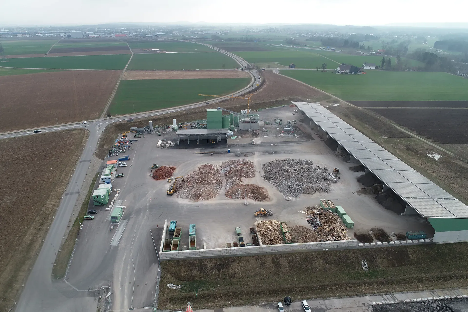 Luftaufnahme einer Abfallwirtschaftsanlage mit sortierten Materialhaufen, Containern und umliegenden Feldern unter einem wolkigen Himmel.