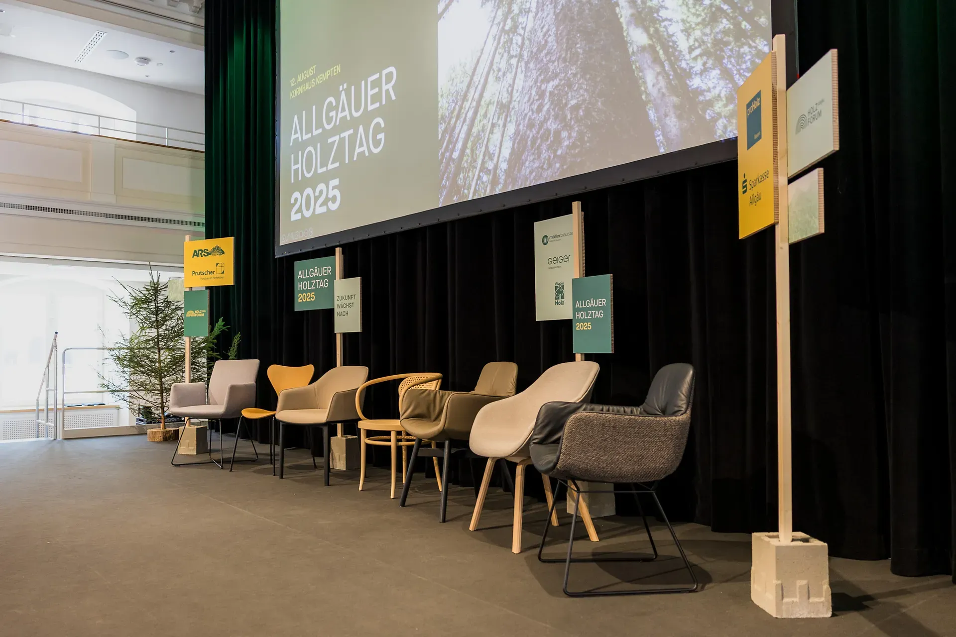 Ein Bühnenaufbau mit sechs Stühlen, einer großen Leinwand mit der Aufschrift „Allgäuer Holztag 2025“ und verschiedenen Schildern, darunter links ein kleiner Baum.