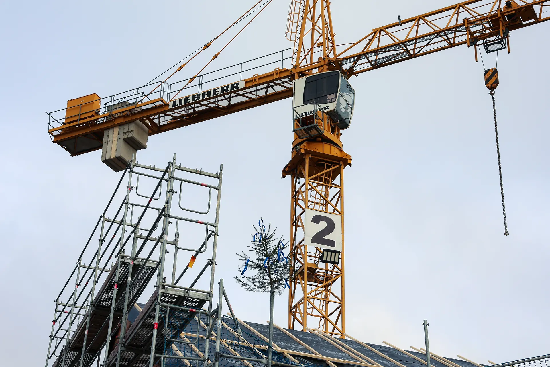 ein Kran über einem Gebäude im Rohbau mit Gerüst und ein kleiner Tannenbaum auf dem Dach mit bunten Bändern