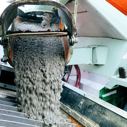 Betonierer lässt frischen Beton auf eine Metalloberfläche fließen, Teil eines Bauprozesses, mit sichtbaren Maschinen im Hintergrund.